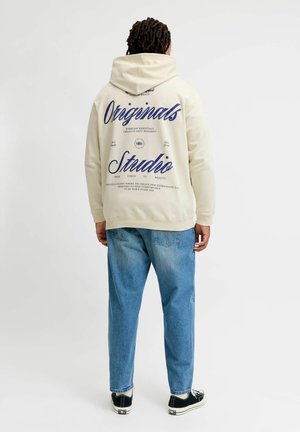 Person mit dunklen lockigen Haaren, die einen cremefarbenen Hoodie mit blauem Text "Originals Studio" und hellblaue Jeans trägt, steht mit dem Rücken zum Betrachter.