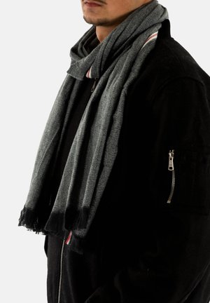 Écharpe grise aux bords effilochés, faite d'un tissu doux, portée autour du cou, avec un subtil accent rayé en rouge et blanc.