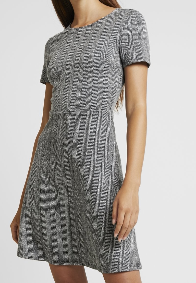 Femme portant une robe grise à motif chevron, manches courtes, longueur au genou, taille cintrée et encolure ronde.