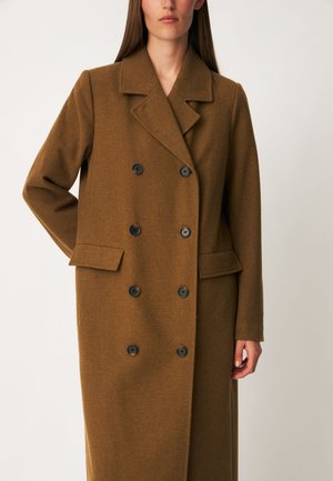 Femme portant un long manteau marron croisé avec des boutons noirs et de larges revers, debout avec un bras derrière le dos.