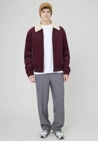 Blouson aviateur en velours côtelé bordeaux avec col en shearling beige, associé à un t-shirt blanc et un pantalon gris à jambes larges, complété par des baskets noires et blanches.