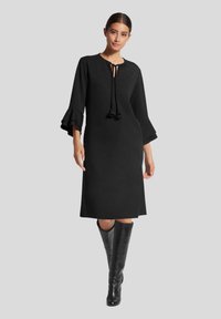 Schwarzes Kleid mit langen Glockenärmeln und einem Ausschnitt mit Schlitz, der mit Bändern versehen ist. Glatter Stoff, knielanges Design und kombiniert mit hohen schwarzen Stiefeln.