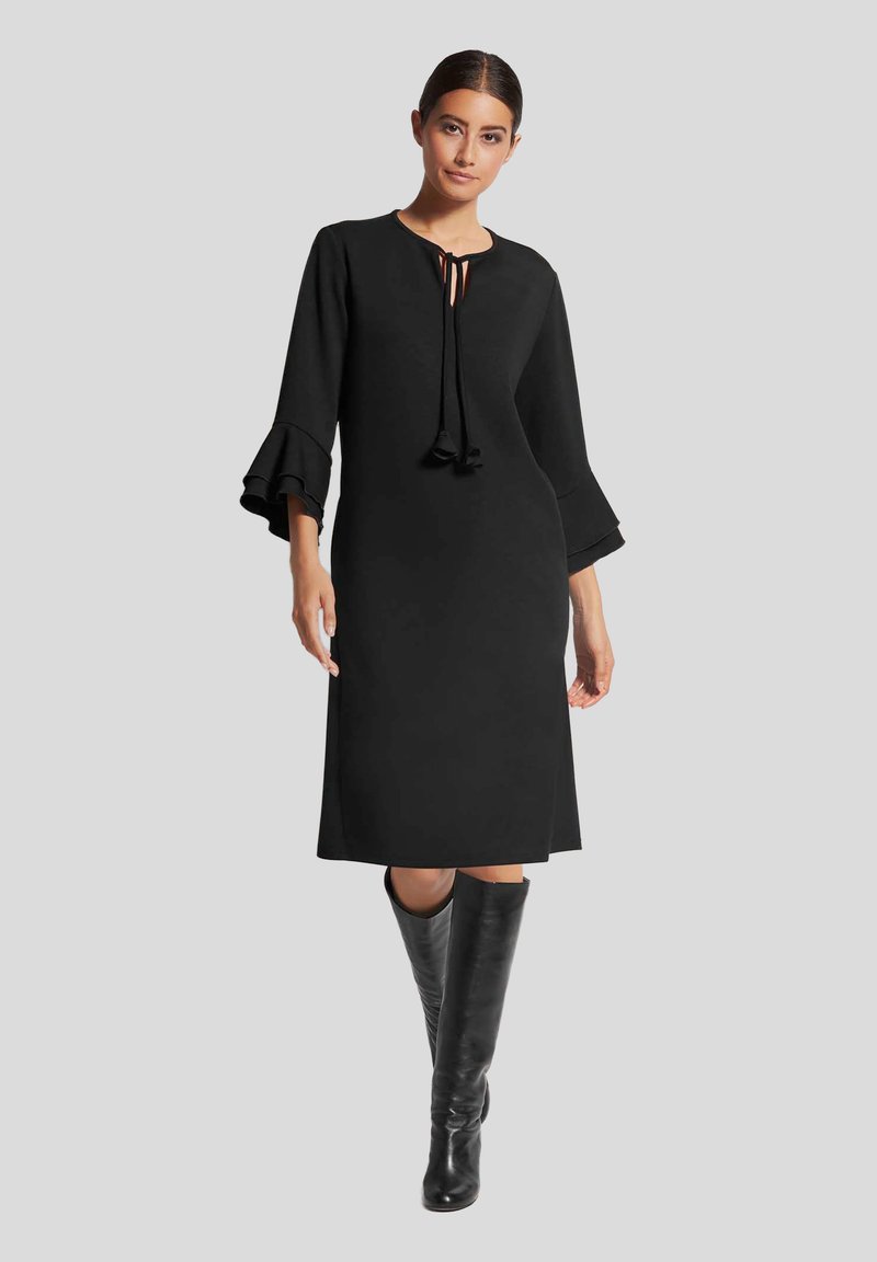 Schwarzes Kleid mit langen Glockenärmeln und einem Ausschnitt mit Schlitz, der mit Bändern versehen ist. Glatter Stoff, knielanges Design und kombiniert mit hohen schwarzen Stiefeln.