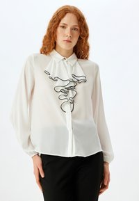 Blusa bianca con colletto a bottoni e ricamo astratto nero sul davanti, realizzata in tessuto leggero e trasparente.