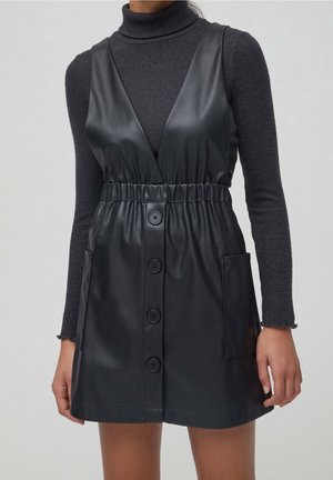 Kobieta ubrana w czarną, bezrękawną sukienkę z ekoskóry z przodu zapinaną na guziki i z kieszeniami, na ciemnoszarym golfie z długim rękawem.