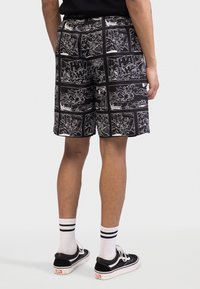 Svarte shorts med tegneserierypremotiv som har hvite design. Materialet virker lett; avsluttet med rett snitt. Brukt med svarte joggesko og stripete sokker.