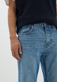 Ljusblå denimjeans med en blekt textur och klassisk femfickorsdesign, bärs med en strukturerad mörkblå stickad väst.