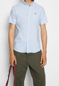 Camicia azzurra a maniche corte con bottoni, colletto rigido, taschino frontale e piccolo logo a corona rossa. Il tessuto sembra liscio e leggero.