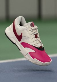 Nike tenisice imaju bijelu gornjište s crvenim naglascima, ružičastu kapu prsta, teksturiranu mrežu i crni obrub. Lagani dizajn s gumiranim potplatom.