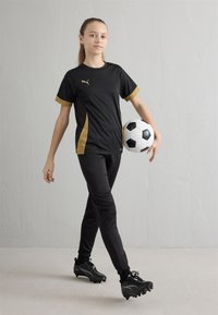 Črna športna majica z zlatimi poudarki, črne hlače in črni kopački, drži črno-belo nogometno žogo, stoji na svetli podlagi.