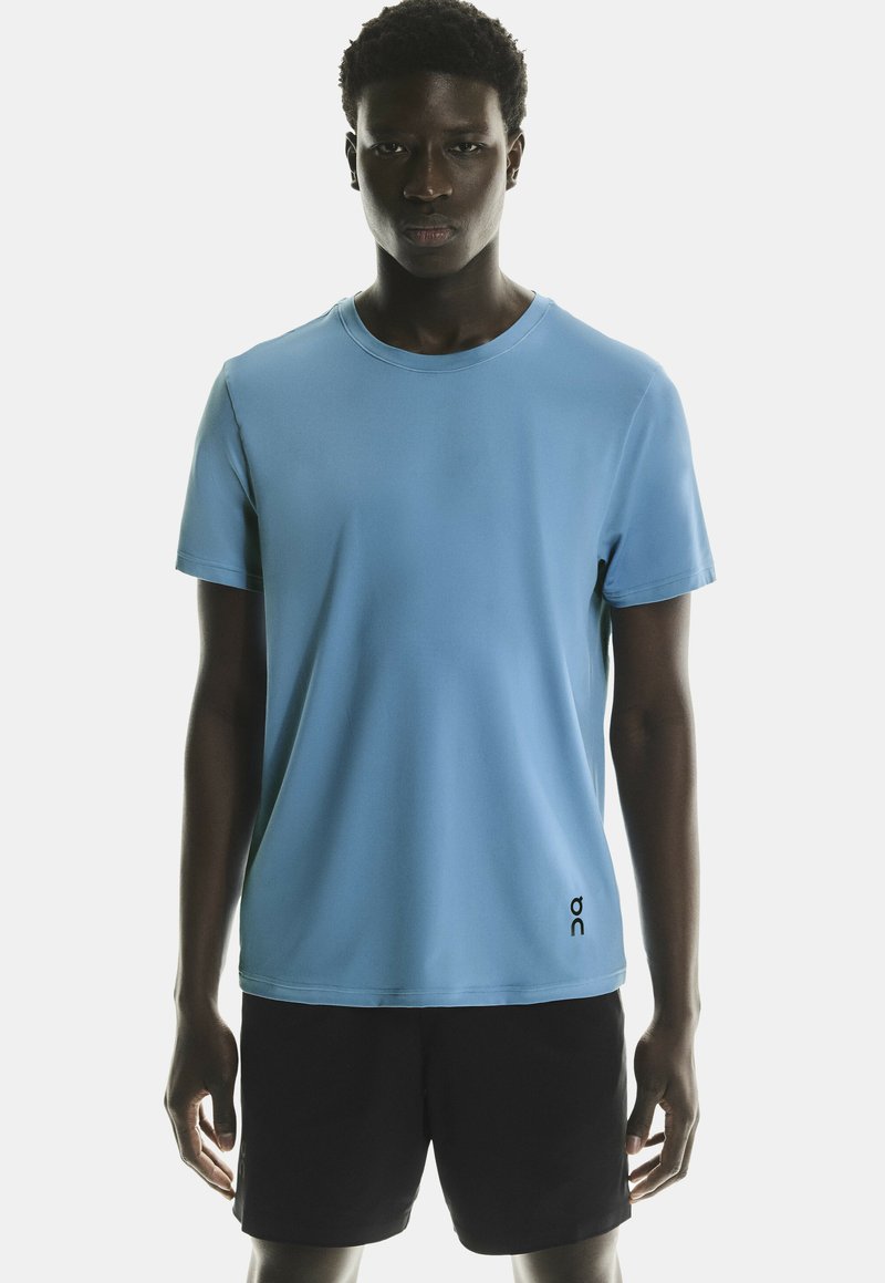T-shirt bleu clair à manches courtes avec un col rond et un détail de logo noir en bas à gauche. Associé avec un short noir.