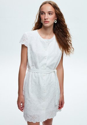 Vestido blanco bordado con mangas cortas de volante, cierre frontal de botones, cinturón de tela y dobladillo en forma de onda, con patrones florales y recortes.
