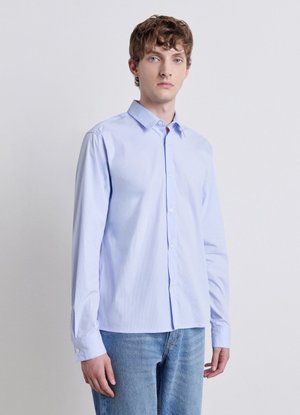 Junger Mann mit kurzem lockigem braunem Haar, der ein hellblaues Hemd mit Knöpfen und blaue Jeans vor einem schlichten Hintergrund trägt.