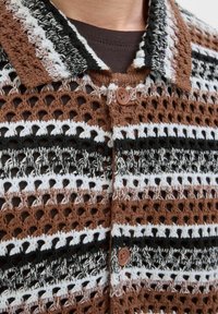 T-shirt en crochet avec un motif rayé brun, noir et blanc, présentant une texture en tricot ajouré et un col à boutons marron.