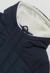 Modra podložena jakna z flis podloženim kapuco, ki ima šivano diamantno vzorec in sprednje zapiranje. Mehka, teksturirana notranjost.