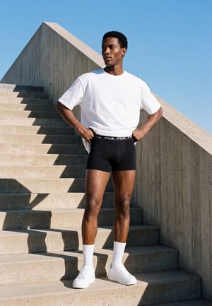 Homme debout sur des escaliers en béton portant un t-shirt blanc, un short noir, des chaussettes blanches et des baskets blanches devant un ciel bleu clair.