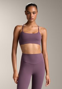 Vijolična športna modrc z tankimi naramnicami, narejena iz gladkega, raztegljivega blaga. Ujemajoče se visoke legice brez šivov dopolnjujejo komplet.