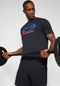 Sort bomulds t-shirt med et blåt og rødt grafiktryk, der står "IRON Paradise." Model løfter en vægtstang iført sorte shorts.