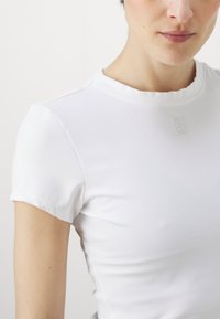 Weißes, eng anliegendes T-Shirt mit kurzen Ärmeln, geripptem Muster und rundem Ausschnitt. Verfügt über ein dezentes, eingeprägtes Logo auf der Brust.