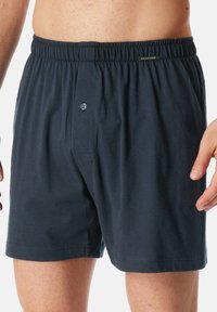 Pantaloni boxer in cotone blu scuro con cintura elastica e un singolo bottone frontale. Texture liscia e design minimalista.