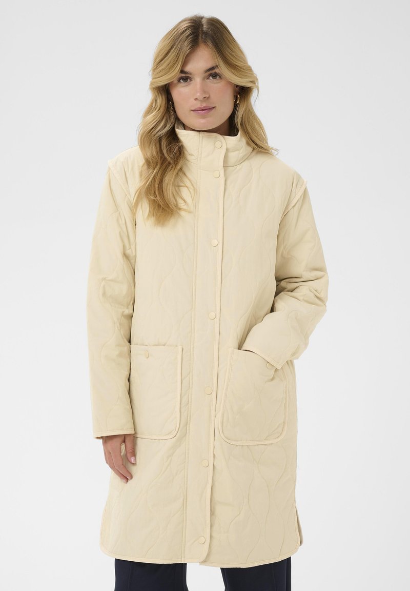 Manteau matelassé beige avec col montant, boutons-pression et deux poches frontales. Texture douce avec un motif subtil en forme de vagues sur le tissu.