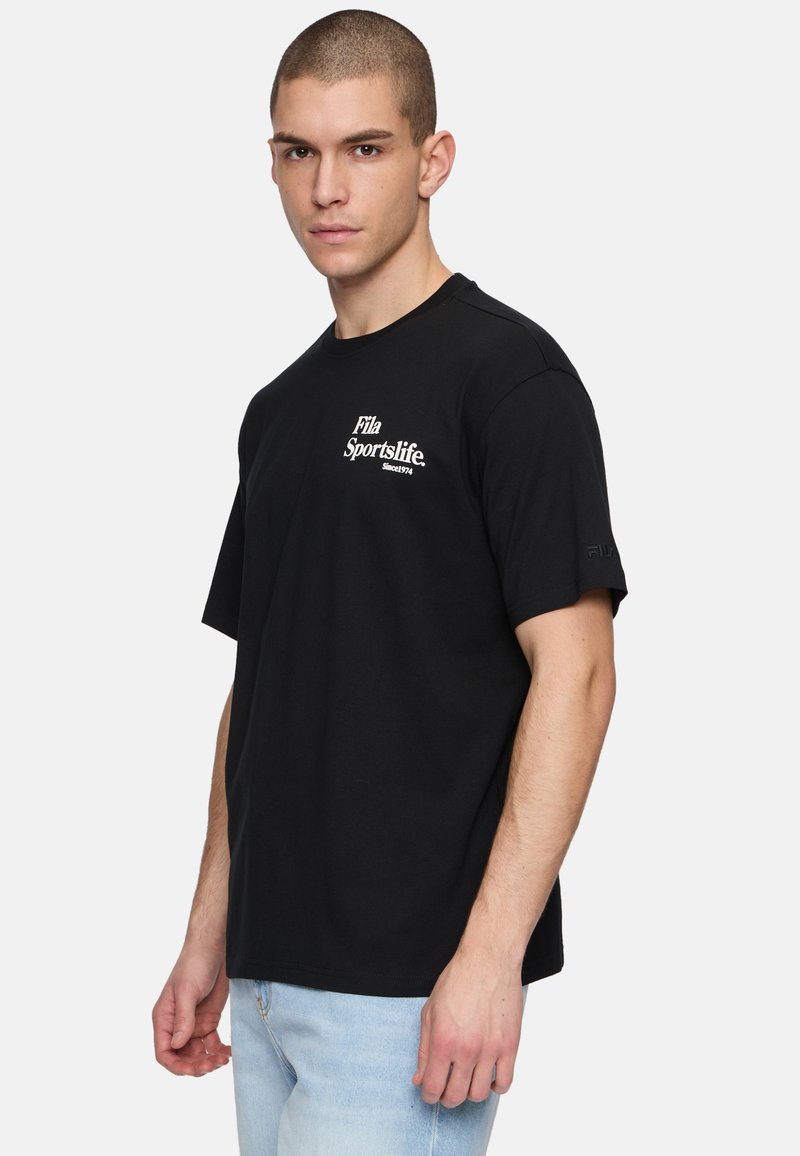 T-shirt negra de manga corta hecha de algodón, que presenta un logo blanco de Fila Sportslife en el pecho y una etiqueta de logo en la manga. Estilizada con jeans de color azul claro.