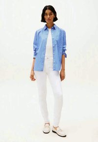 Μπλε και άσπρη ριγέ πουκάμισο με κουμπιά, συνοδευόμενο από άσπρο τοπάκι και άσπρα τζιν. Μοντέλο με άσπρα loafers, που στέκεται μπροστά σε ουδέτερο φόντο.