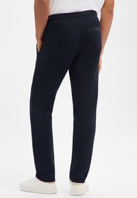 Mężczyzna ubrany w czarne, zwężane spodnie z kieszeniami po bokach i z tyłu, białe tenisówki oraz białą koszulę, stoi na jednolitym białym tle.