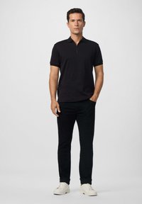 Muž stojaci v čiernom polo tričku s krátkym rukávom, čiernych nohaviciach a bielych teniskách, na jednoduchom svetlom pozadí.