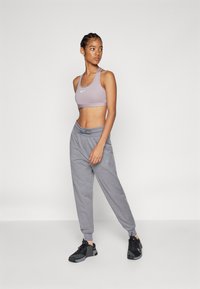 Sutiã desportivo lilás claro com um logo branco, combinado com calças de fato cinzentas em corte tapered com cintura ajustável. Um ténis desportivo preto completa o visual.