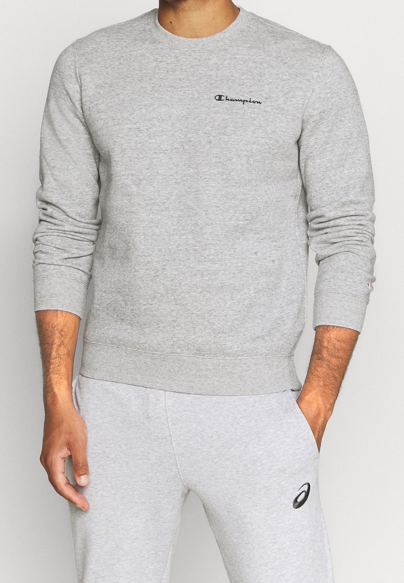 Homme portant un sweat-shirt Champion gris clair et un pantalon de survêtement Asics gris clair, debout avec une main dans la poche.