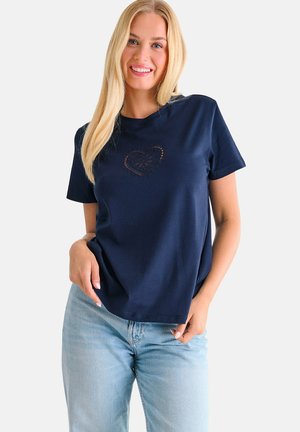 Femme souriante aux longs cheveux blonds portant un t-shirt bleu marine avec un motif de cœur et un jean bleu clair, posant devant un fond uni.