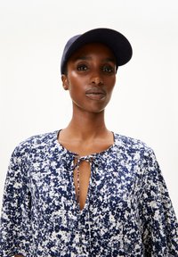 Navy- und weiß gemustertes Kleid mit einem Schlüsselloch-Ausschnitt und Bindedetail. Der Stoff ist leicht und hat eine weiche Textur. Eine passende Kappe rundet das Outfit ab.