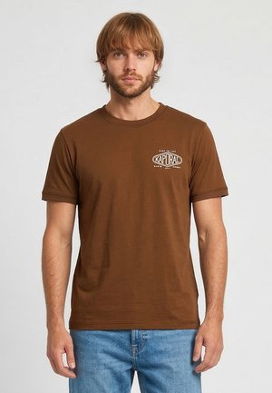 Man met lichtbruin haar en baard, draagt een bruin Kaporal t-shirt en blauwe spijkerbroek, staand tegen een effen lichte achtergrond.