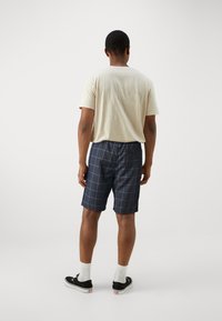 Camiseta de cuello redondo beige combinada con pantalones cortos navy que presentan un patrón de cuadrícula en azul claro. Zapatillas negras y calcetines blancos completan el look.