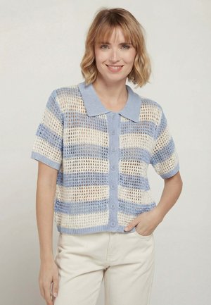 Femme aux cheveux blonds courts portant un haut en maille à manches courtes boutonné, avec des rayures horizontales bleues et blanches, posant avec une main dans la poche.