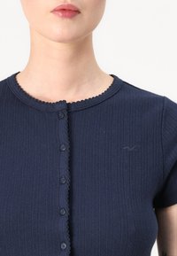 Haut à manches courtes côtelé bleu marine avec un col en dents de scie, fermeture à cinq boutons et un détail de logo subtil sur le côté gauche.