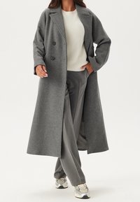 Manteau gris en laine mélangée à double boutonnage, coupe décontractée, col à revers crantés et poches latérales, porté sur un pull crème et un pantalon gris.