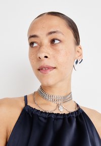 Cercei în formă de inimă din argint, colier din argint cu strasuri, top halter din satin bleumarin cu decolteu plisat, piele netedă cu pistrui.