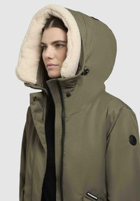 Olijfgroene geïsoleerde jas met een pluche beige capuchonvoering, ritssluiting, zijzakken en een logo-opdruk op de mouw. Glad oppervlak.