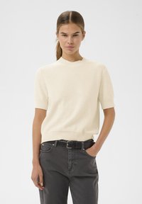 Svijetlosmeđa pletena majica s kratkim rukavima i okruglim izrezom, u kombinaciji s tamnosivim hlačama. Suptilna tekstura, ležeran kroj.