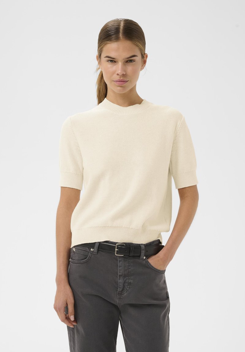 Svijetlosmeđa pletena majica s kratkim rukavima i okruglim izrezom, u kombinaciji s tamnosivim hlačama. Suptilna tekstura, ležeran kroj.