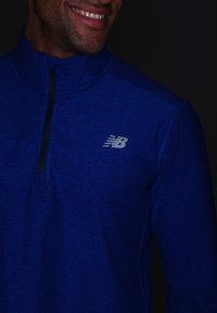 Blå atletisk pullover i melerat material, med en halvdragkedja och grå "NB"-logotyp på vänster bröst. Mjuk, stretchig tyg.