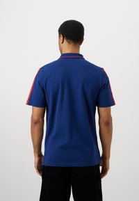 Μπλε πόλο μπλούζα με κόκκινες λεπτομέρειες στους ώμους, κοντά μανίκια και γιακά. Φτιαγμένη από υφή, ελαστικό ύφασμα.