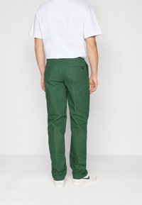 Green cotton trousers with a straight-leg design, two back pockets, and a subtle brand tag near the waistband. Worn with a white t-shirt.