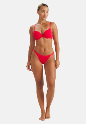 Frau steht barfuß und trägt ein passendes rotes BH- und Tanga-Set, blickt leicht nach links vor einem schlichten weißen Hintergrund.