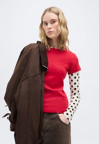 Νεαρή γυναίκα με ξανθά σγουρά μαλλιά, φορώντας κόκκινο τοπ με κοντά μανίκια πάνω από λευκό μακρυμάνικο με μαύρες πουά, κρατώντας σκούρο καφέ σακάκι.