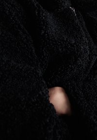 Černá fleecová bunda s texturovaným, chlupatým povrchem; má zip, částečně otevřený, odhalující ruku.