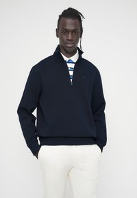 Πουλόβερ ναυτικού μπλε με φερμουάρ στο ένα τέταρτο, με ριμπ μανσέτες και τελείωμα, σε συνδυασμό με λευκό και μπλε ριγέ πόλο πουκάμισο από κάτω. Λείο ύφασμα.