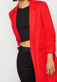Blazer rojo brillante con un diseño estructurado, que presenta un solo botón y una tela texturizada, usado sobre un top negro corto y pantalones oscuros.